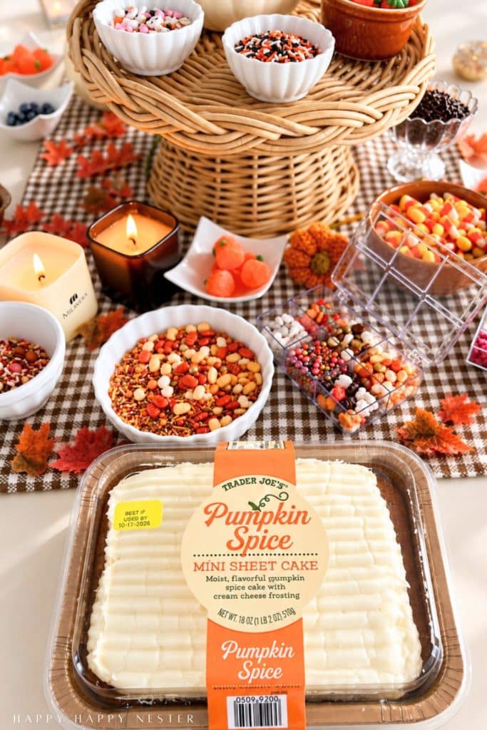 Packaged Trader Joe's pumpkin spice mini sheet cake surrounded by bowls of Halloween and fall sprinkles, candy pumpkins, and decorating supplies on a gingham tablecloth.
