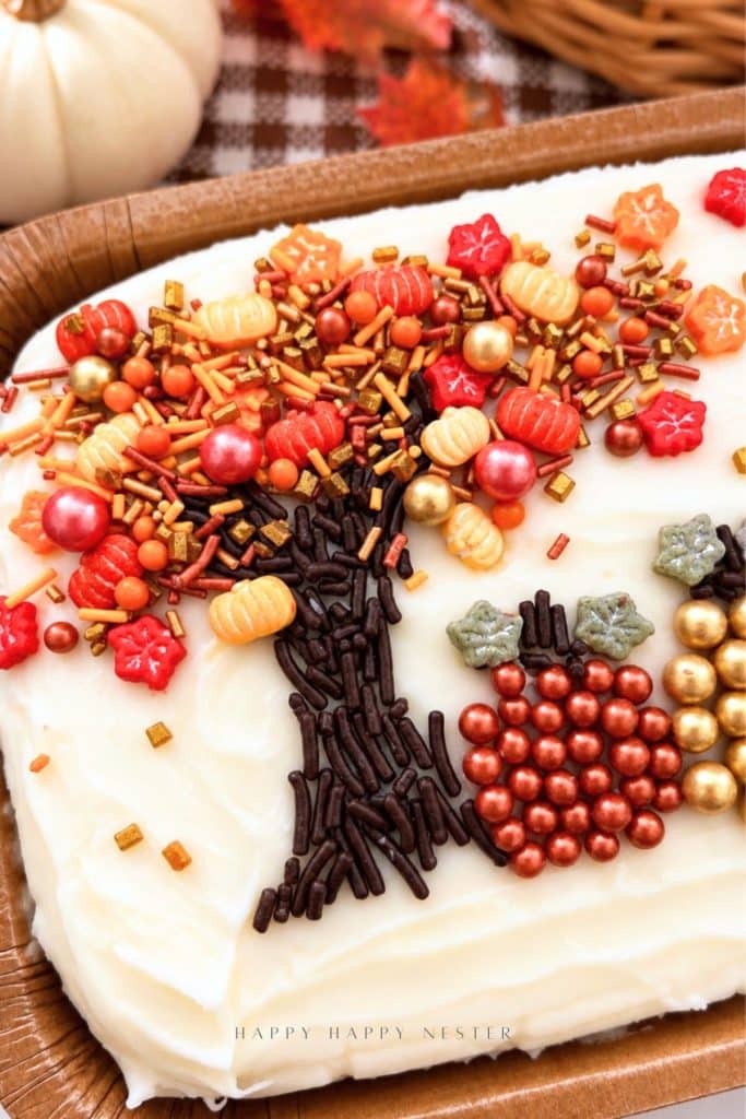 Close-up of a fall cake decorated with a chocolate sprinkle tree, orange and gold sprinkles, candy leaves, and pumpkins made from metallic sugar pearls.
