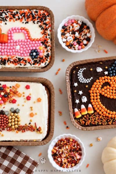 Three decorated mini sheet cakes featuring a fall sprinkle tree with pumpkins, a pink candy truck carrying pumpkins, and an orange jack-o'-lantern on chocolate frosting.