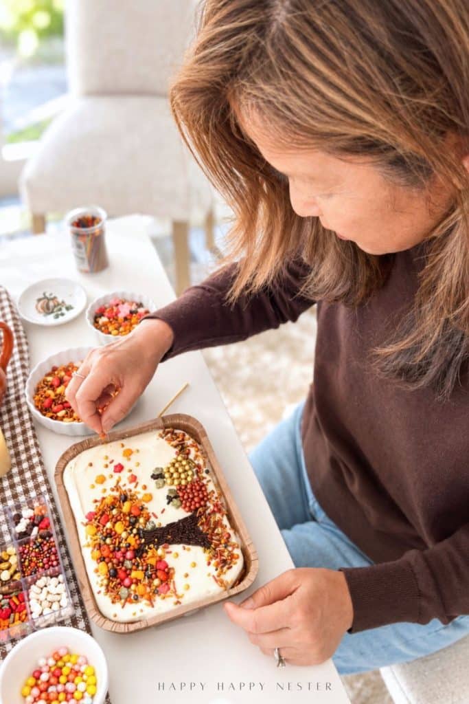 Woman decorating a Trader Joe's mini sheet cake with colorful fall sprinkles, a chocolate sprinkle tree, and candy pumpkins at a table filled with decorating supplies.