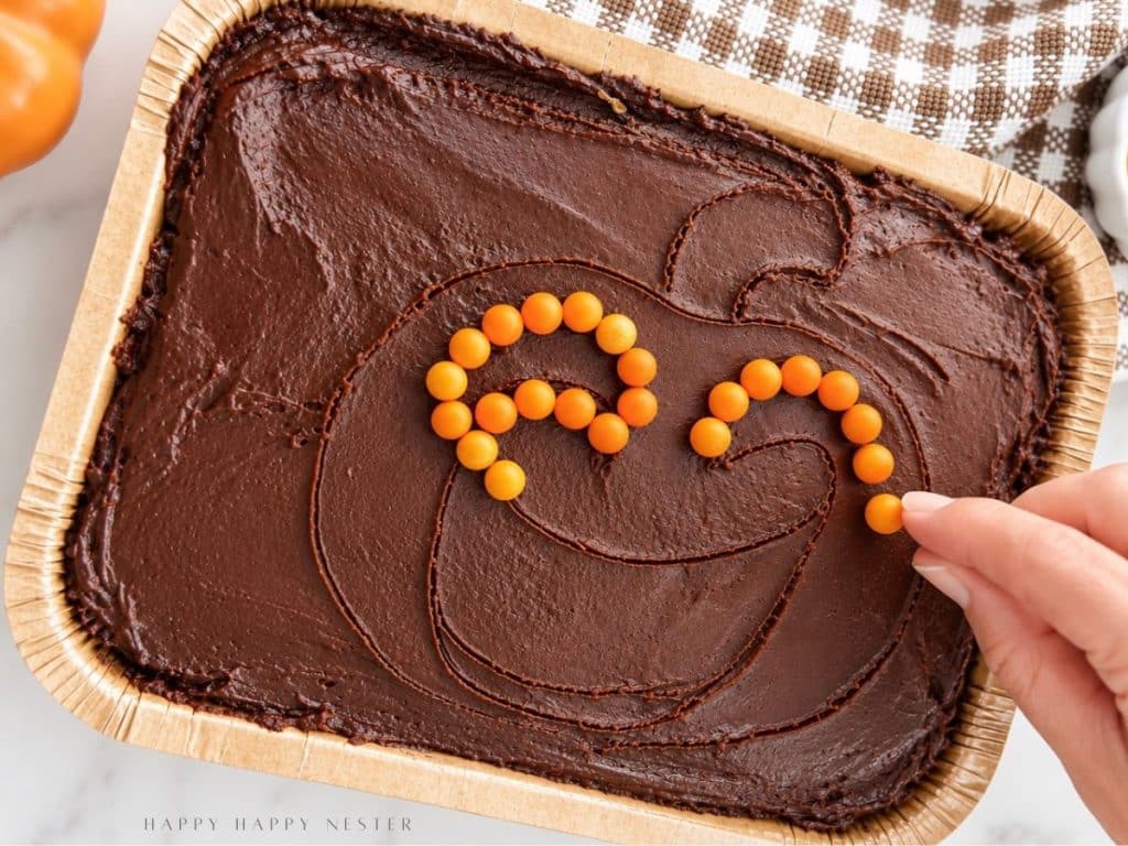 Close-up of a hand placing orange candy pearls along a jack-o'-lantern face outline on a chocolate-frosted mini sheet cake.